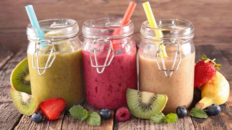 Three colorful smoothies in jars for toddlers: avocado green, strawberry pink, and banana yellow, served on a wooden table.