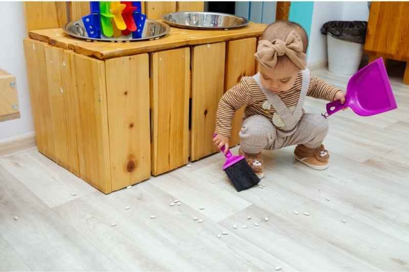 A little baby girl sweeping the floor with a toy broom and dustpan as part of a Montessori practical life activity. 