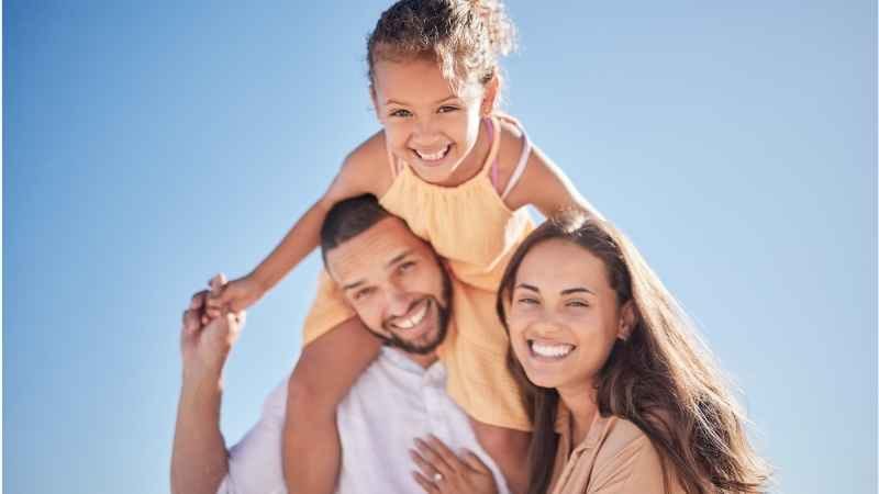 Smiling father carrying daughter on his shoulders with mother by his side, representing positive parenting and calm family moments.