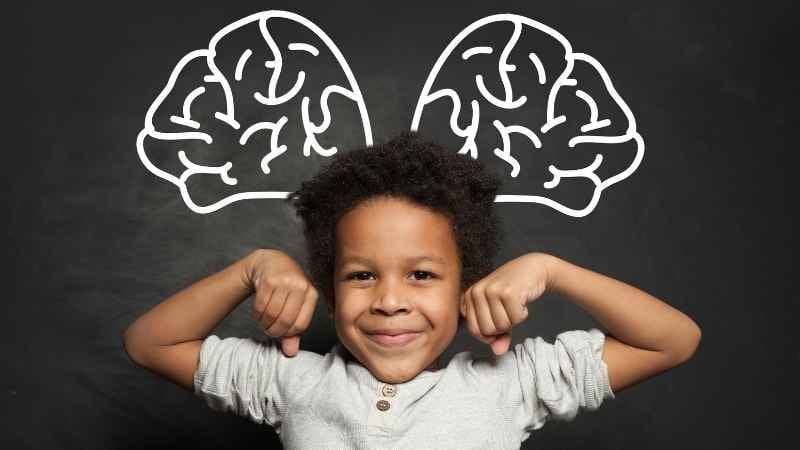 A toddler raising both arms confidently with a brain drawing sketched in white chalk on a blackboard behind, symbolizing child brain development.