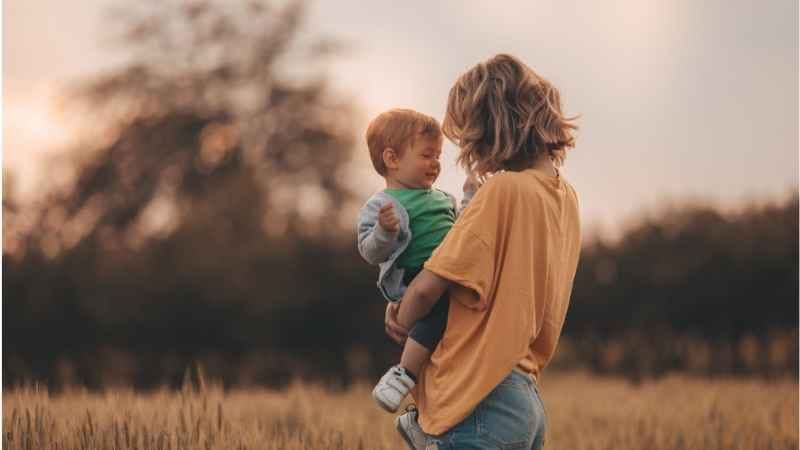 Mother standing in a field on a late afternoon holding her son in her lap, symbolizing authentic motherhood and the unfiltered beauty of parenthood.