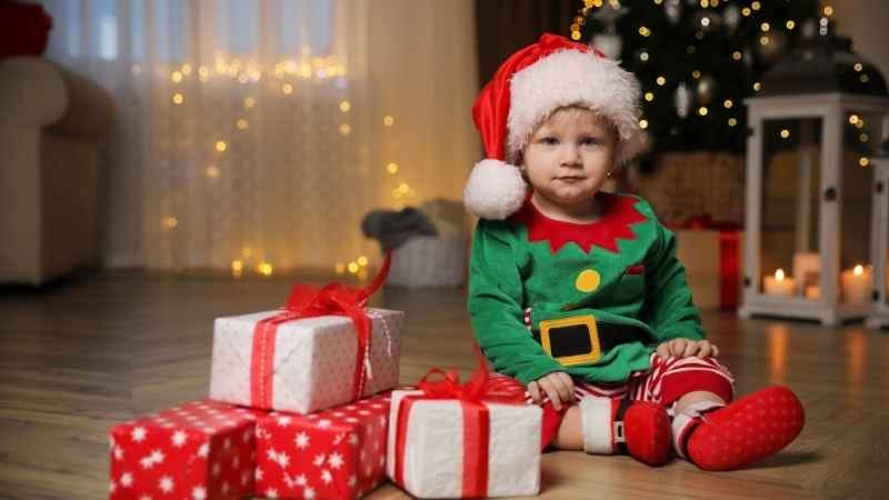 Baby boy Christmas outfit sitting on the floor behind Christmas decorations with gifts around him, wearing a festive green & red romper and holiday clothes.
