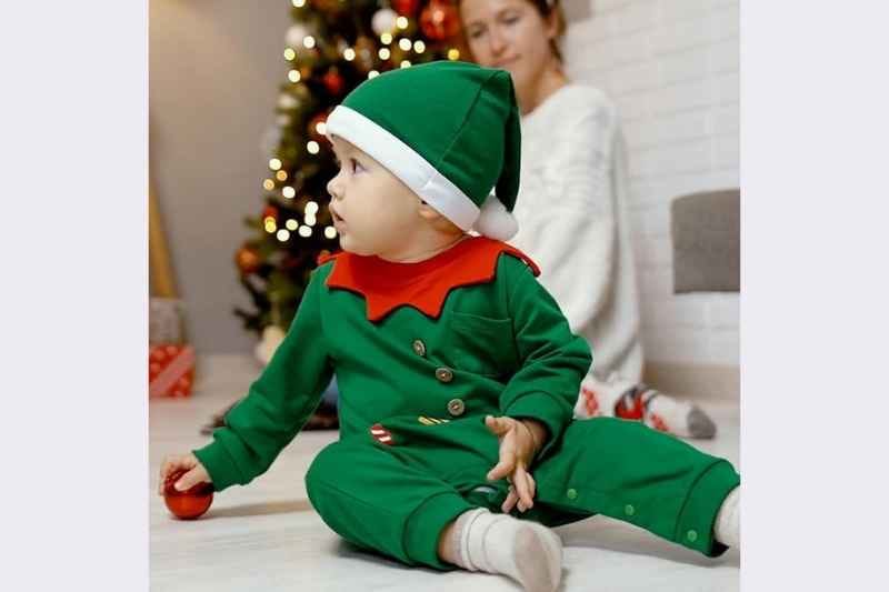 Baby boy standing on the floor wearing the Hifot My First Christmas Baby Romper & Hat Set in green, soft festive romper with matching hat; blurred mother and Christmas decorations in the background; perfect newborn and infant Christmas outfit for photoshoots, holiday gatherings, and first Christmas memories.