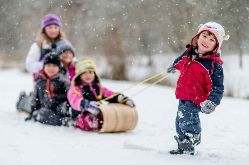 A toddler sliding joyfully down a snowy hill on a sled, laughing and enjoying outdoor winter play, with a parent watching nearby.
